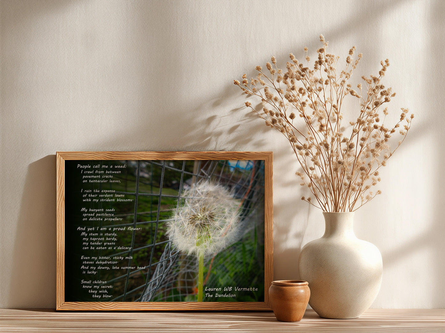 Framed artwork with a dandelion and text next to a vase with dried flowers on a shelf.