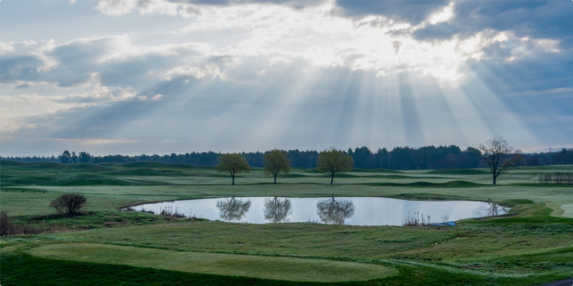 Sun beams break through dramatic clouds over a small reflecting pond.