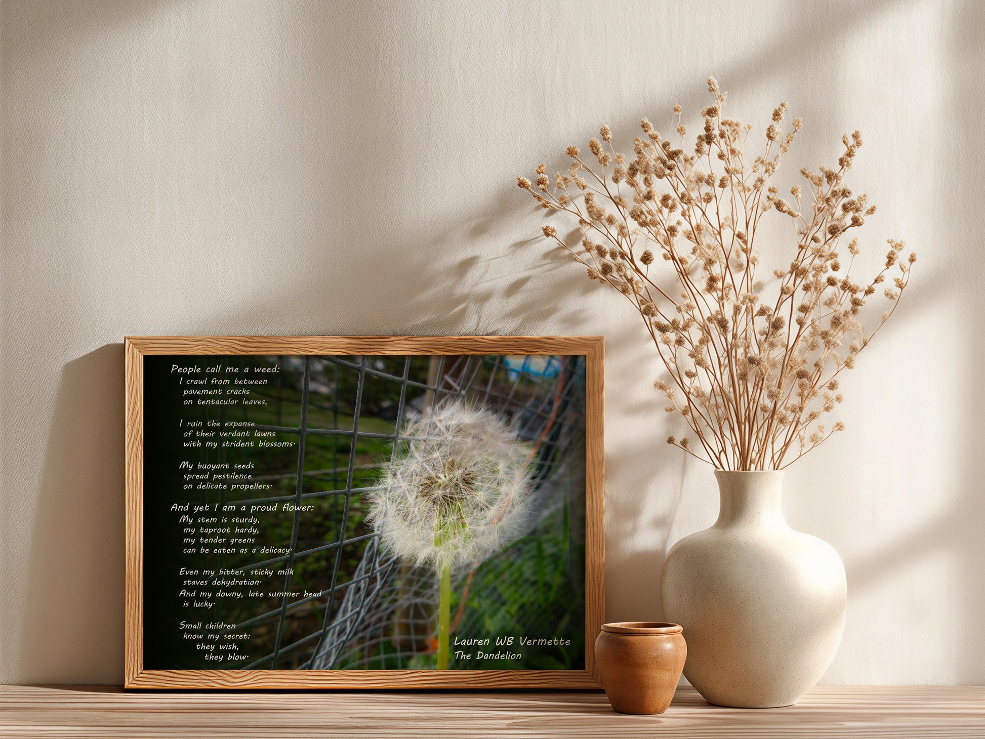 Framed artwork with a dandelion and text next to a vase with dried flowers on a shelf.