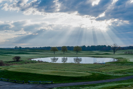 Sun beams break through dramatic clouds over a small reflecting pond.